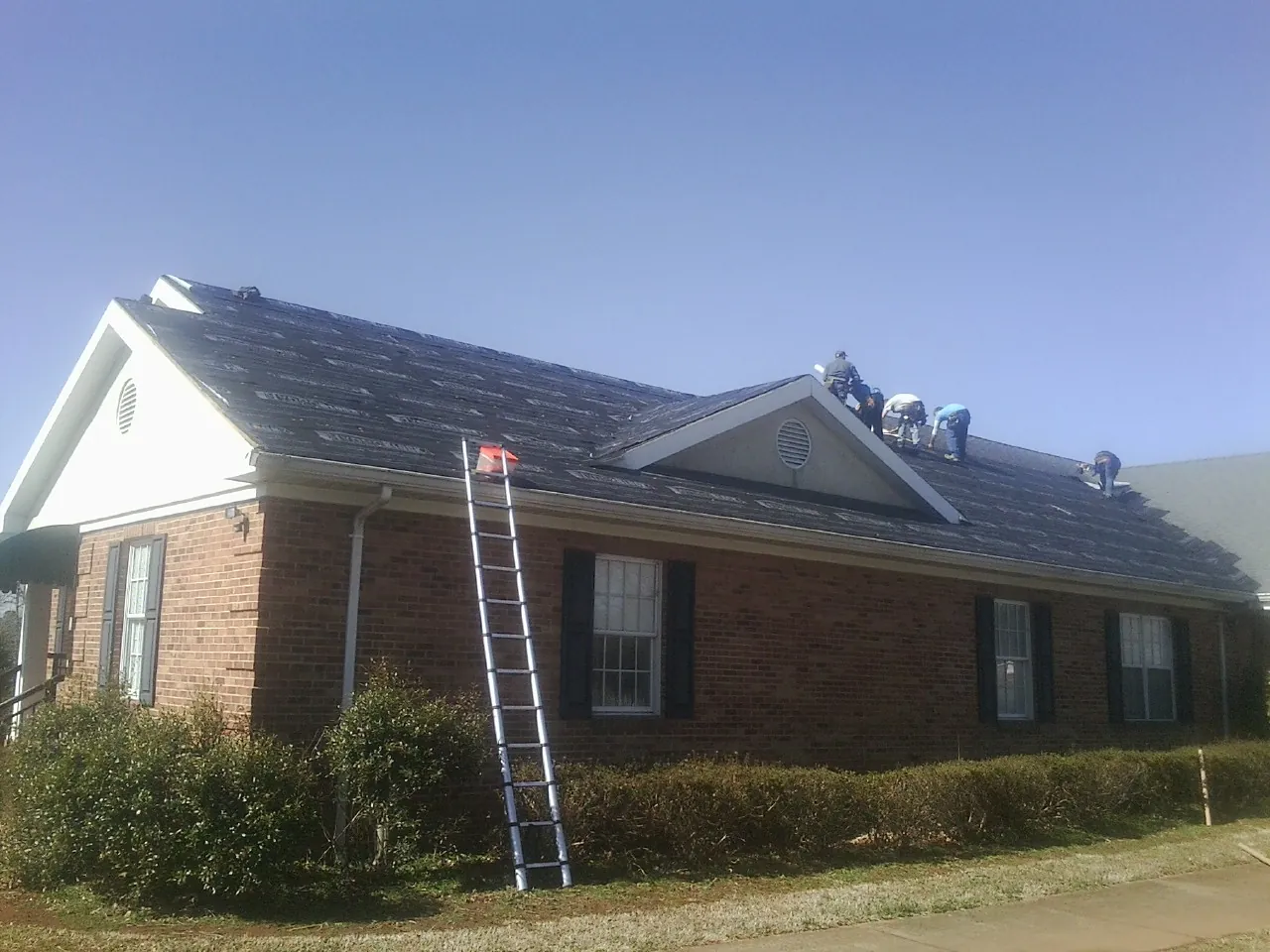 Crew tearing shingles off a brick ranch roof with a ladder set — Emergency Roof Repair work by our crew in Waxhaw