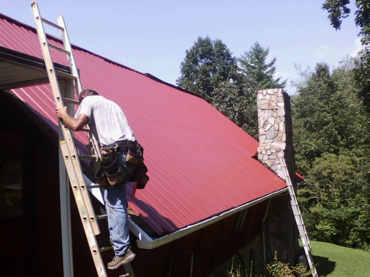 Roofer on a ladder beside a red metal roof and stone chimney — Soffit & Fascia Repair by Roofing Brothers in Waxhaw